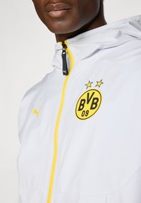Homme portant une veste blanche de Borussia Dortmund avec une fermeture éclair jaune, logo BVB 09 et logo Puma, gros plan sur le haut du torse et le visage.