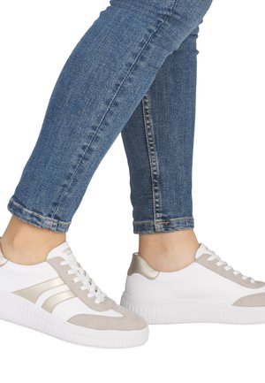 Fødder iført hvide og beige sneakers kombineret med blå denimjeans, vist mod en hvid baggrund.
