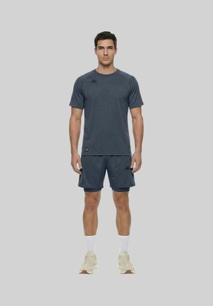 Uomo in piedi, volto in avanti, indossa abbigliamento sportivo grigio scuro, calzini bianchi e sneakers beige su sfondo grigio uniforme.