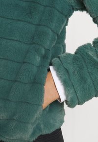 Teal faux-fur topp med en texturerad, ribbad design. Ärmar och nederkant är mjuka; plagget har en avslappnad passform med en synlig vit skjorta under.