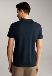 Dunkelblaues Poloshirt mit kurzen Ärmeln, strukturierter Musterung und Kragen, von hinten getragen von einem männlichen Model, kombiniert mit beigen Shorts.
