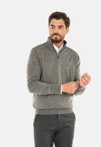 Maglione grigio con zip realizzato in materiale morbido, con polsini e orlo a costine, indossato sopra una camicia bianca con colletto.