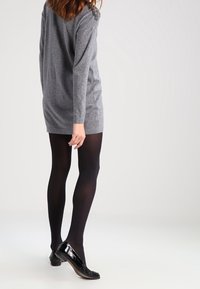 Abito lungo grigio a maniche lunghe con orlo arrotondato, indossato sopra collant neri e scarpe nere lucide con tacco; presenta dettagli decorativi sulle spalle.