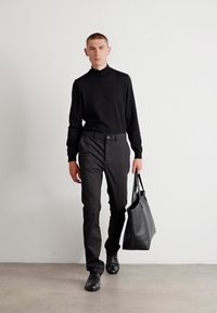 Zwarte coltrui, fitted zwarte broek en zwarte schoenen. Het model draagt een stijlvolle zwarte leren tote-bag. Neutrale achtergrond.