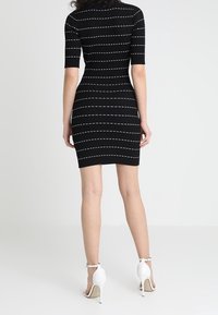Abito nero lavorato a maglia con maniche corte e righe orizzontali bianche, forma aderente, texture minimale. Indossato con sandali bianchi con tacco alto.