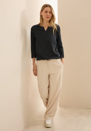 Zwarte V-hals lange mouwen top gecombineerd met beige, broek met elastische taille. Model staat tegen een neutrale achtergrond en draagt witte sneakers.