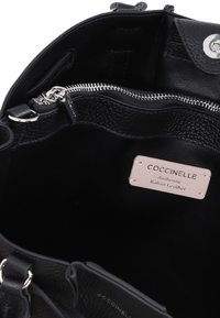 Interno di una borsa nera in pelle con una tasca con zip, texture liscia e un'etichetta rosa chiaro con la scritta "Coccinelle Authentic Italian Leather."