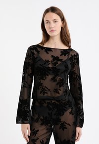 Woman wearing a black sheer top and pants with velvet floral patterns, standing against a plain white background.