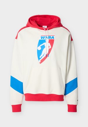 Junger Mann trägt einen weißen, roten und blauen WNBA-Hoodie und marineblaue Basketballshorts, steht vor einem schlichten weißen Hintergrund.