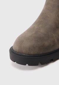 Bruine matte leren laarsneus met dikke zwarte rubberen profielzool op een witte achtergrond, die textuur en zoolprofieldetails toont.
