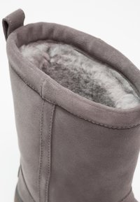 Botte en daim gris avec doublure intérieure en fourrure grise pelucheuse et languette en tissu à l'arrière de l'ouverture de la cheville.