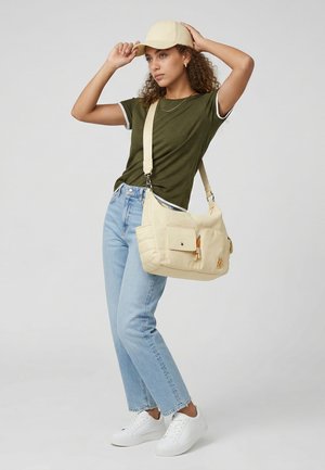 Mujer con camiseta verde, vaqueros azul claro, zapatillas blancas, gorra beige y llevando un bolso grande beige de hombro con múltiples bolsillos.