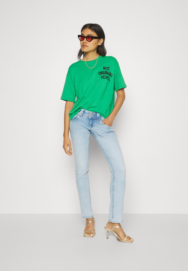 Grünes, übergroßes T-Shirt mit dem Aufdruck "NOT ORDINARY PEOPLE", kombiniert mit hellblauen Skinny Jeans und metallischen, strapazierfähigen High Heels. Schwarze Sonnenbrille getragen.