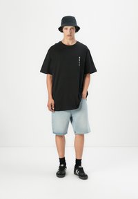Schwarz T-Shirt mit kurzen Ärmeln und subtiler Grafik auf der linken Seite, hellblaue Denim-Shorts, schwarzer Bucket Hat und schwarze Schuhe mit weißen Streifen.