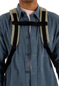 Cinghie nere regolabili si attaccano a un'imbragatura imbottita grigia. Indossato sopra una camicia blu con texture, con chiusura a bottoni e taschino sul petto.