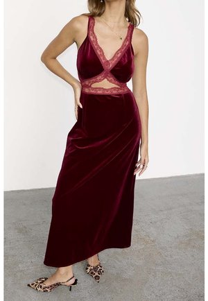 Femme portant une robe sans manches en velours bordeaux avec des découpes en dentelle et des escarpins à bride arrière à imprimé léopard, debout sur un sol gris.