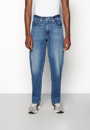 Pantalones de mezclilla azul claro con corte recto, acabado desgastado, dos bolsillos delanteros y cierre de botón. Llevados con zapatillas deportivas grises.
