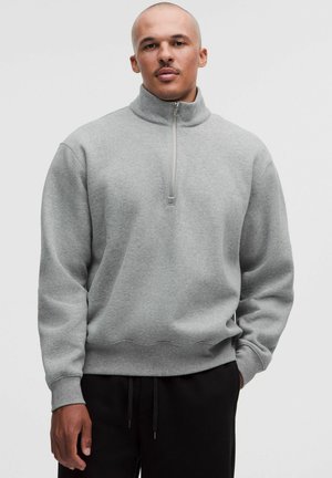 Mann mit rasiertem Kopf, der einen hellgrauen Pullover mit Viertel-Reißverschluss und schwarze Hose trägt, steht vor einem einfarbigen hellen Hintergrund.