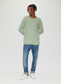 Groene gebreide trui, slim-fit blauwe spijkerbroek en witte sneakers met zwarte accenten. De trui heeft een ronde halslijn en lange mouwen.