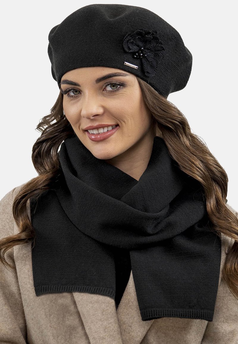 Sort strikket beret med blomsterdetalje, parret med et matchende sort tørklæde. Begge har en glat tekstur og minimalistisk design.