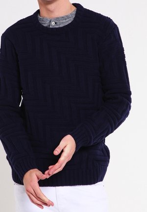 Man wearing a textured dark navy sweater over a gray henley shirt, paired with white pants, standing against a plain white background.