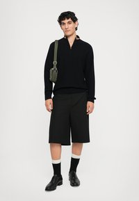 Uomo che indossa un maglione nero con zip, pantaloni corti neri fino al ginocchio, scarpe nere con calze bianche e una borsa a tracolla verde oliva, in piedi su uno sfondo bianco.