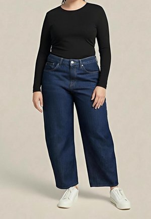 Osoba ubrana w ciemnoniebieskie jeansy o szerokich nogawkach, czarną bluzkę z długim rękawem i białe wsuwane trampki, stojąca na jednolitym tle.