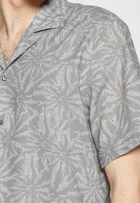 Homme portant une chemise grise à manches courtes, boutonnée, avec un motif floral abstrait et un col ouvert, laissant apparaître une partie de la poitrine et du bras.