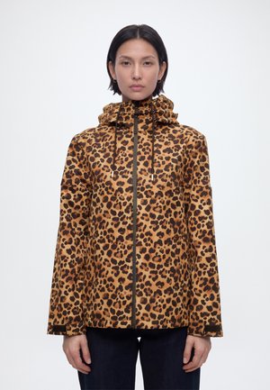 Nő barna és fekete leopárdmintás kapucnis dzsekiben, elöl cipzárral és húzózsinórokkal, sima fehér háttér előtt állva.