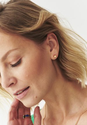 Gold round stud earring worn on the left ear, hair partially covering the ear. Smooth texture, minimalist design, with natural skin tones visible.