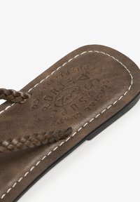 Gros plan sur la semelle d'une sandale en cuir marron avec des bandes tressées, avec des coutures blanches et un texte de marque embossé sur la semelle intérieure.