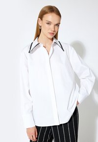 Camisa blanca con un cuello festoneado negro en contraste, mangas largas y cierre de botones en el frente. Se lleva con pantalones a rayas en blanco y negro.