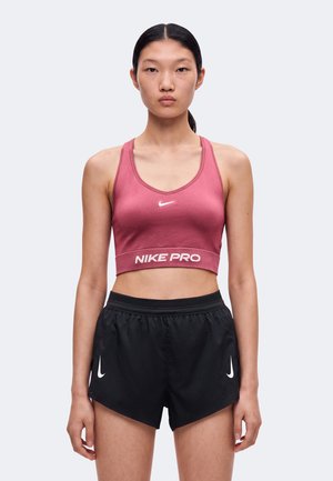 Γυναίκα φοράει ένα ροζ Nike Pro crop top χωρίς μανίκια και μαύρο αθλητικό σορτς Nike, στέκεται μπροστά σε απλό φόντο.