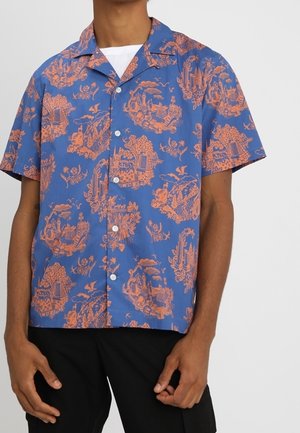 Hombre vistiendo una camisa azul de manga corta con botones y patrones escénicos y florales intrincados en naranja, combinada con pantalones negros.