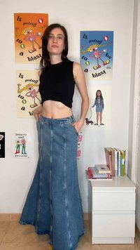 Γυναίκα με μαύρο αμάνικο crop top και μακριά καμπάνα τζιν φούστα που στέκεται σε ένα δωμάτιο με πολύχρωμες αφίσες και ένα λευκό συρταριέρα με βιβλία.