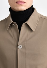 Beige jakke med tekstureret stof, med knappelukning foran og spids krave. Bæres over en sort ribbet turtleneck.