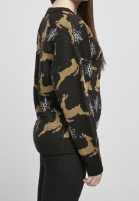 Frau in einem schwarzen Pullover mit goldenen Rentier- und weißen Schneeflockenmuster, kombiniert mit schwarzen Hosen, seitlich gezeigt.