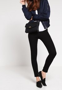 Donna che indossa una bomber jacket blu navy, una maglietta bianca, pantaloni skinny neri, mocassini neri e tiene una borsa a tracolla nera, in piedi su uno sfondo bianco.