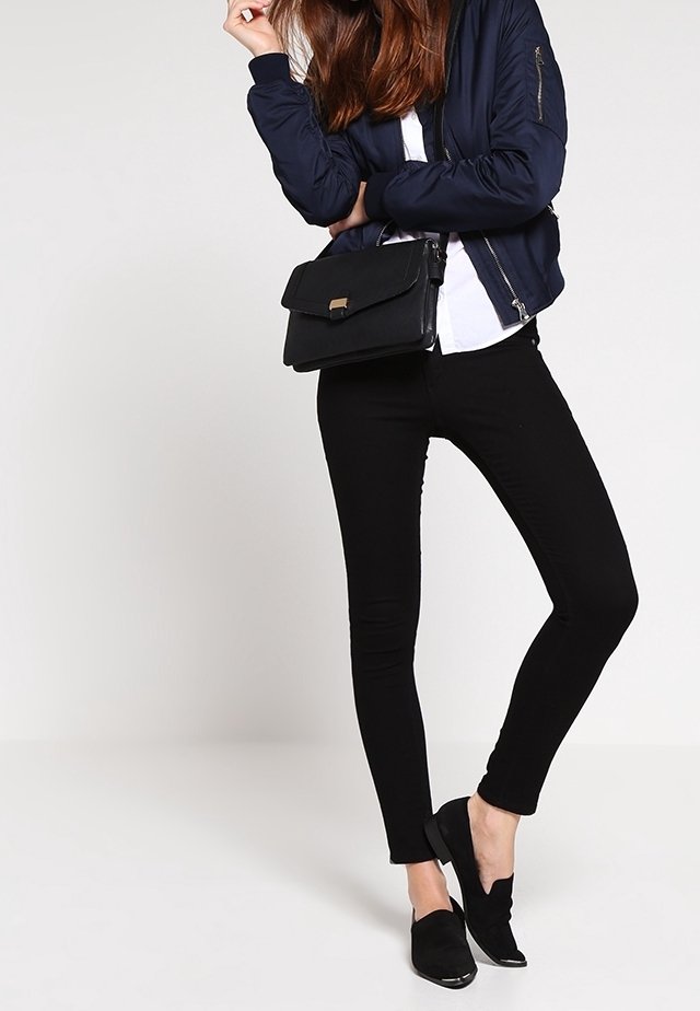 Donna che indossa una bomber jacket blu navy, una maglietta bianca, pantaloni skinny neri, mocassini neri e tiene una borsa a tracolla nera, in piedi su uno sfondo bianco.