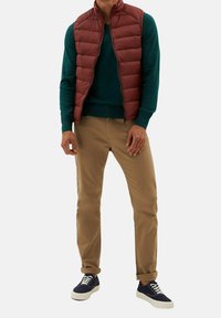 Gepolsterte kastanienbraune Weste über einem türkisfarbenen Pullover, kombiniert mit khakifarbenen Hosen und marineblauen Sneakers. Das Outfit verbindet Lagen und neutrale Töne.