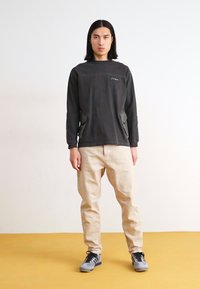 Maglietta nera a maniche lunghe con tasche laterali e finitura testurizzata, abbinata a pantaloni beige chiaro e sneakers grigie, in piedi su un pavimento giallo.