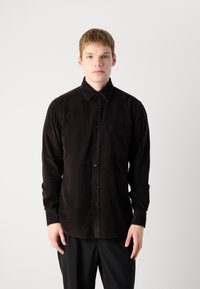 Young man wearing a long-sleeve black corduroy button-up shirt with front pocket, standing against a plain light background.