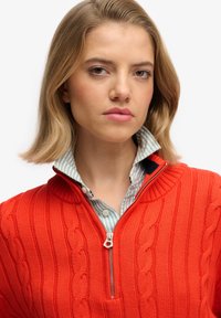 Pull en tricot rouge avec col zippé, présentant des motifs en torsades. Porté par-dessus une chemise à rayures bleu clair et blanche. Texture douce.