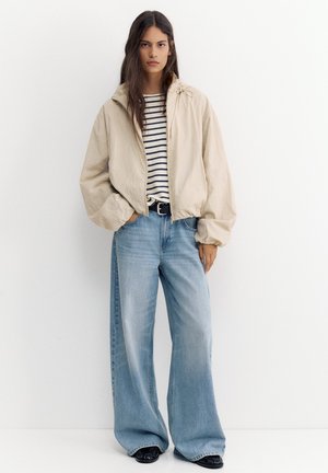 Femme aux longs cheveux foncés portant une veste beige, une chemise rayée, un jean large bleu clair, une ceinture noire et des chaussures noires, debout contre un mur blanc.