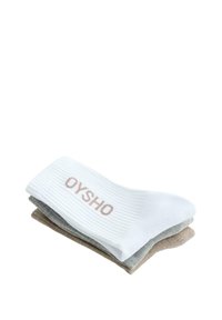Trois chaussettes côtelées pliées en blanc, gris et beige, avec le texte "OYSHO" sur le bord-côte de la chaussette blanche.