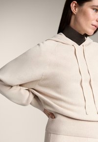 Bežová rebrovaná mikina s kapucňou, s nastaviteľnou šnúrkou, kontrastnou textúrou na pleciach a priliehavým spodným okrajom. Nosená cez tmavý rolák.