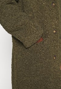 Mørkegrøn strikket uldsfrakke med skildpaddeskal knapper og en sidelomme, der holder en hånd.