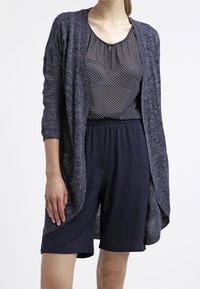 Långärmad, öppen cardigan i mörkgrått stickat tyg, lager på lager över en marinblå topp med prickar och matchande marinblå shorts.