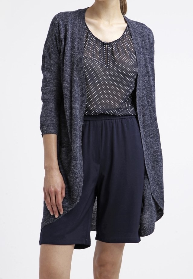 Långärmad, öppen cardigan i mörkgrått stickat tyg, lager på lager över en marinblå topp med prickar och matchande marinblå shorts.