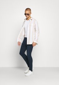 Joven vestido con gafas de sol, camiseta blanca, camisa de rayas abierta, jeans oscuros ajustados y zapatillas blancas, de pie contra un fondo claro y liso.